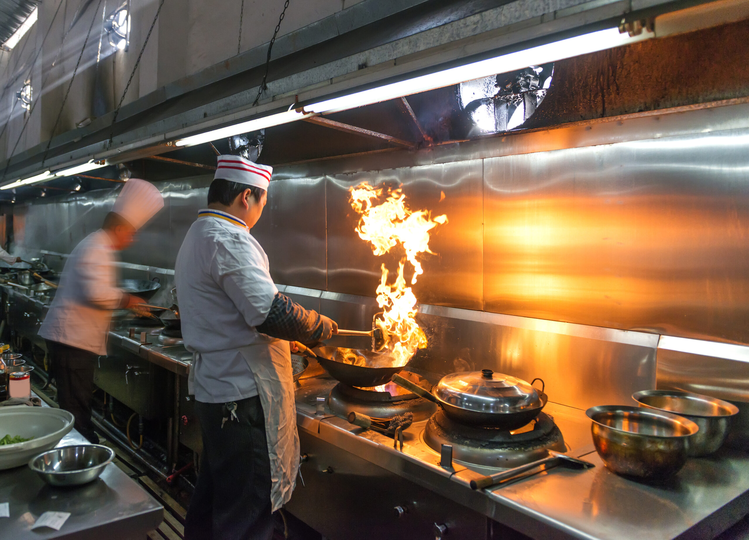 Chef cooking in a commercial kitchen with flames rising from a wok, highlighting the importance of kitchen exhaust systems for fire safety and grease management.